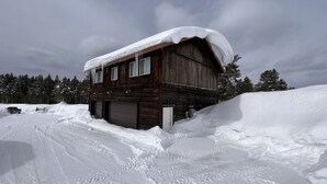 Une cabane en bois dont le toit est recouvert de neige, entourée d’un paysage enneigé et d’arbres couverts de neige.