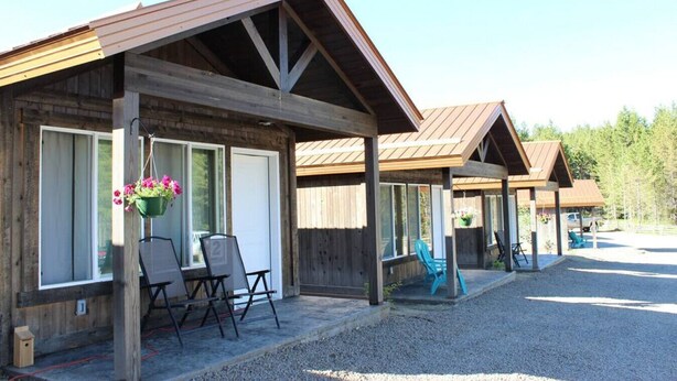 A row of wooden cabins with attached porches, each featuring a table and chairs, and flower baskets hanging from the porch posts. - Shared Hot Tub, WIFI, 2 Queen Beds, Full Bathroom (Island Park)