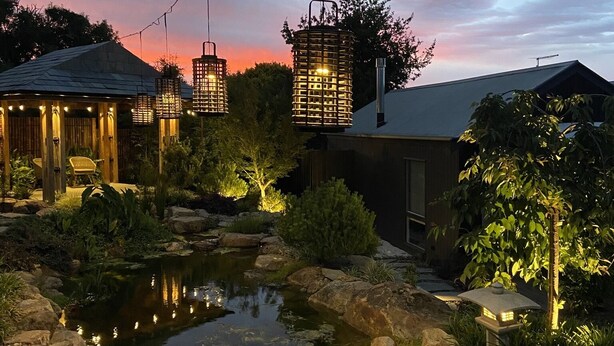 Ein friedlicher Garten mit Teich, umgeben von Steinen und Pflanzen, beleuchtet mit Lichterketten.