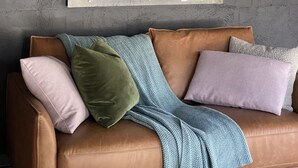 Ein gerahmtes Kunstwerk mit einer menschlichen Figur an einer strukturierten Wand, ein braunes Ledersofa mit Kissen und eine blaue Strickdecke.