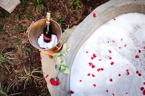 Una bottiglia di champagne in un secchio, una vasca da bagno con petali di rosa e una rosa su una superficie in pietra.