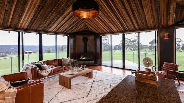 A spacious living room with a large fireplace, leather sofas, and a view of a green landscape through large windows. - Unique luxury + salvage + African theming = Heaven (Hepburn Springs)