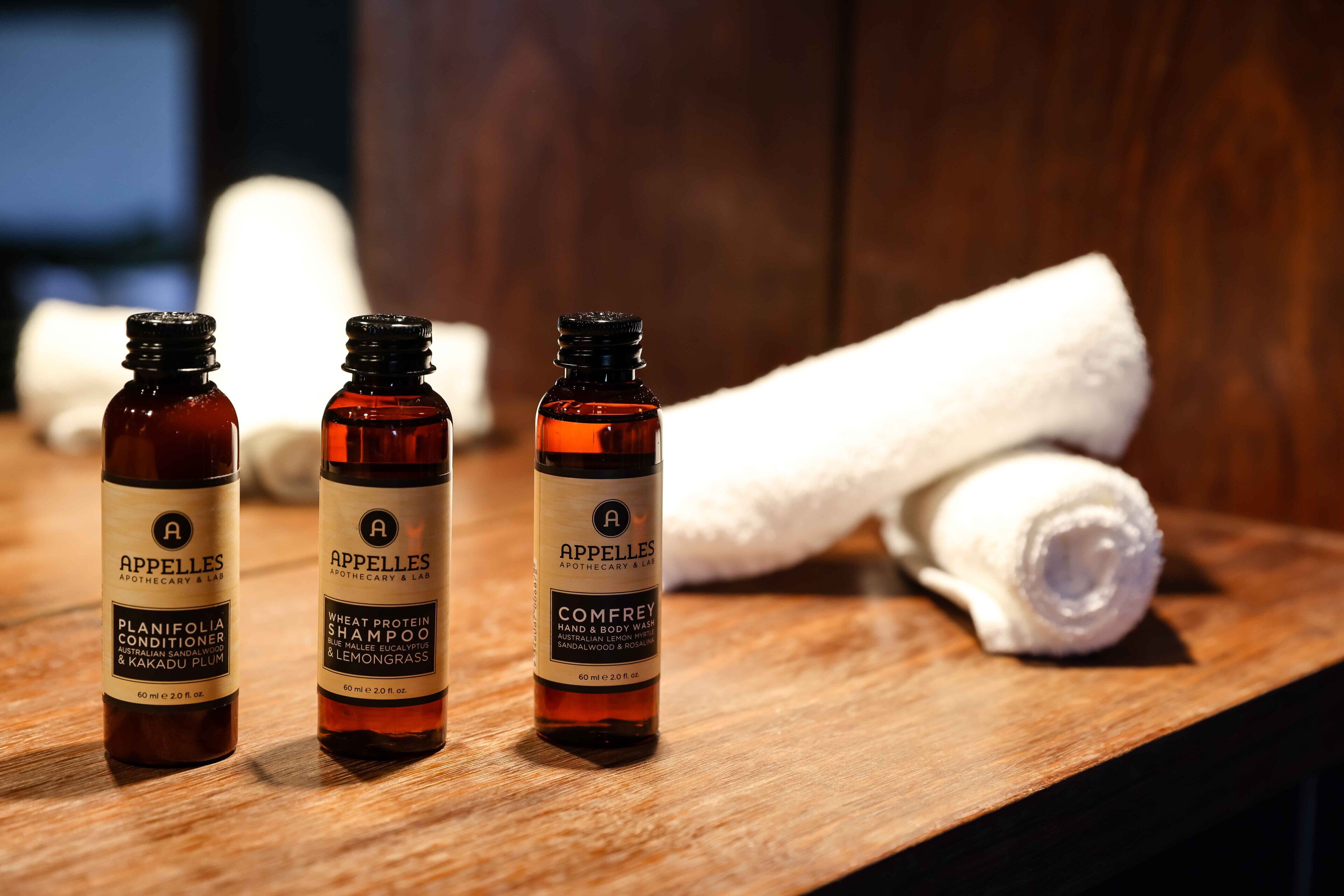 Three bottles of APPELLLES body wash on a wooden surface with a rolled white towel in the background.