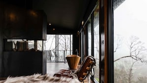 Un interno moderno con un pouf rivestito in pelliccia, una ciotola di legno e una poltrona in pelle vicino a una grande finestra con vista su un paesaggio innevato.