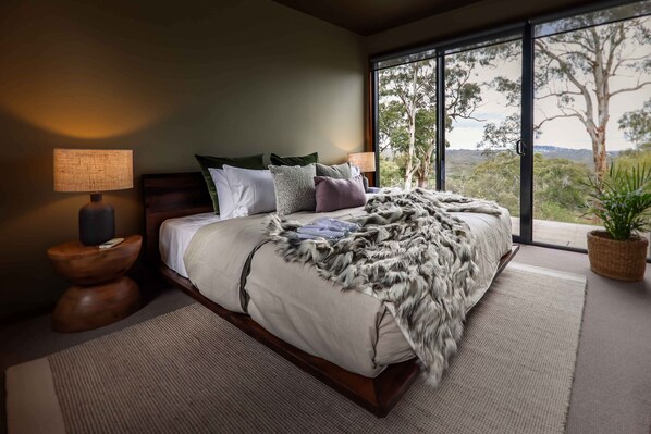 A bedroom with a large bed, a nightstand, a lamp, and a view of trees through a window.