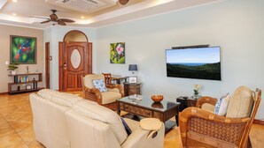 A spacious living room with a large flat-screen TV, comfortable seating, a ceiling fan, and a wooden door. - Mariners Point C1 – 3-Bedroom Villa on Bluff Above Playa Hermosa with Private Pool (Playa Hermosa)