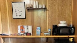 A small kitchenette with a microwave, coffee maker, and a trash bin. There is a bed with a striped bedspread and a framed quote on the wall.