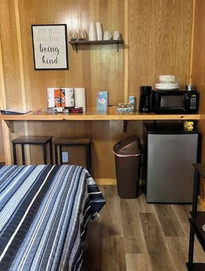 A small kitchenette with a microwave, coffee maker, and a trash bin. There is a bed with a striped bedspread and a framed quote on the wall.