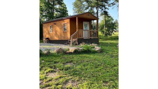 A wooden cabin with a porch and steps leading to a door, surrounded by grass and trees. - Studio Cabin – Pet-Friendly Retreat Near Lake Greeson & Kirby Landing Marina (Kirby)