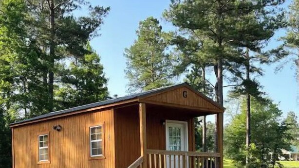 A wooden cabin with a porch and steps leading to a door, surrounded by grass and trees.