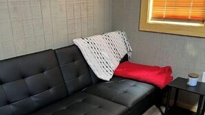 Un sofá de cuero negro con una manta blanca y negra a rayas y una almohada roja. Una mesita de noche negra con una taza blanca está junto al sofá.