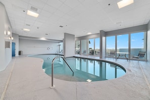 Uma piscina interior ampla com design curvo, ladeada por um chão em mosaico e uma parede de vidro.