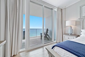 A hotel room with a balcony, a bed with a blue blanket, a nightstand with a lamp, and a view of the ocean.