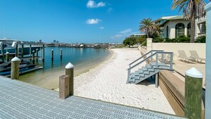 Una playa con una escalera de madera que lleva a un área de descanso, un puerto deportivo con barcos y un edificio con una palmera.
