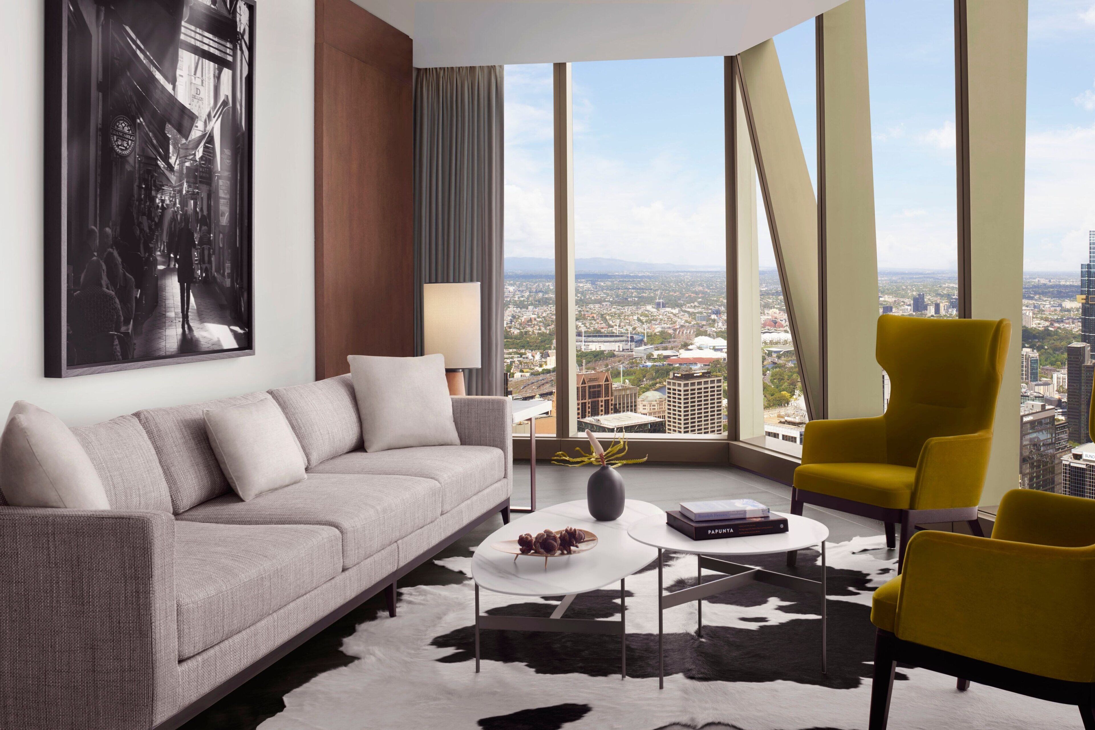 A modern living room with a sofa, a coffee table, and two yellow chairs. A large window offers a city view.