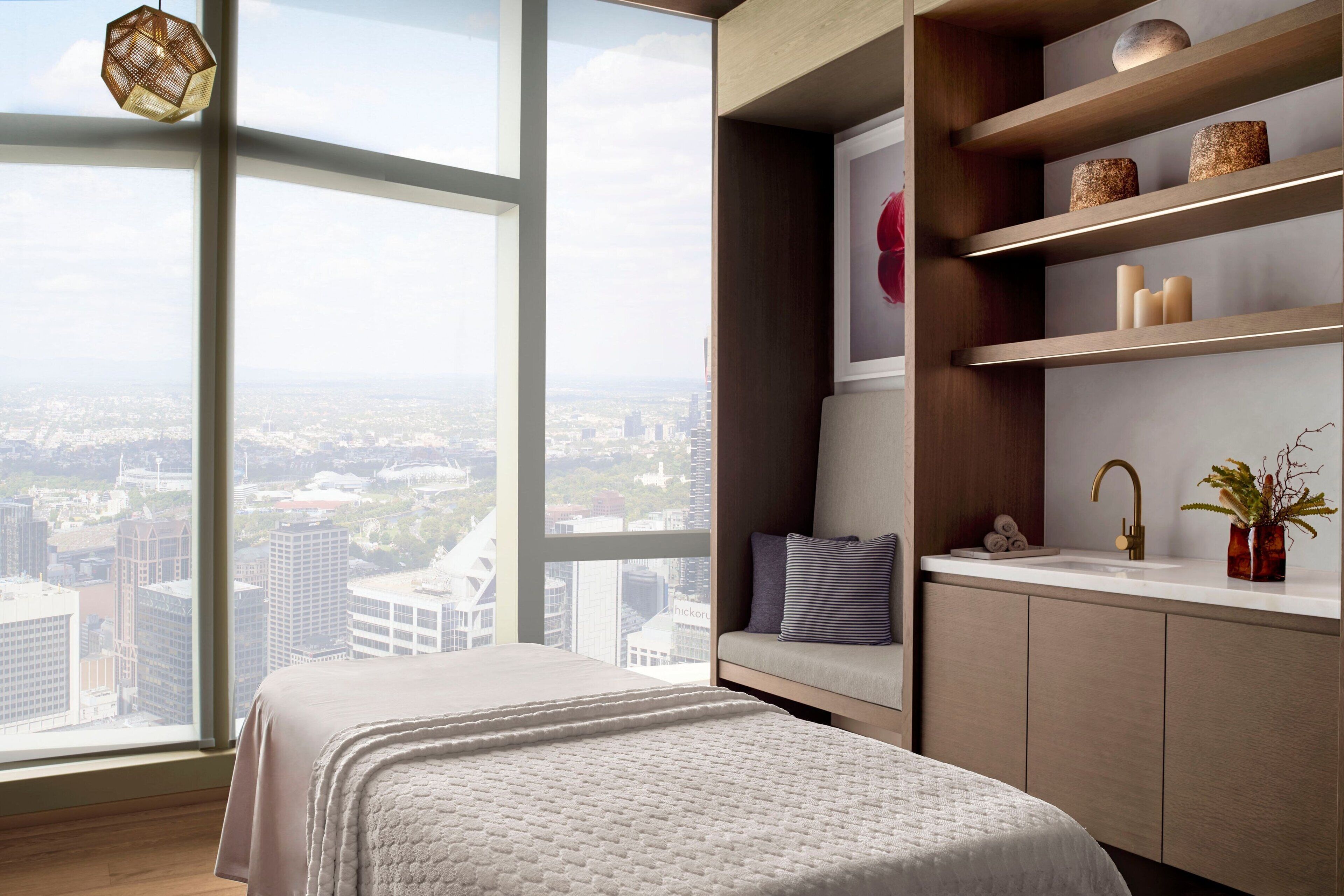 A modern bedroom with a large window, a bed, a built-in shelf with decorative items, and a view of the city.