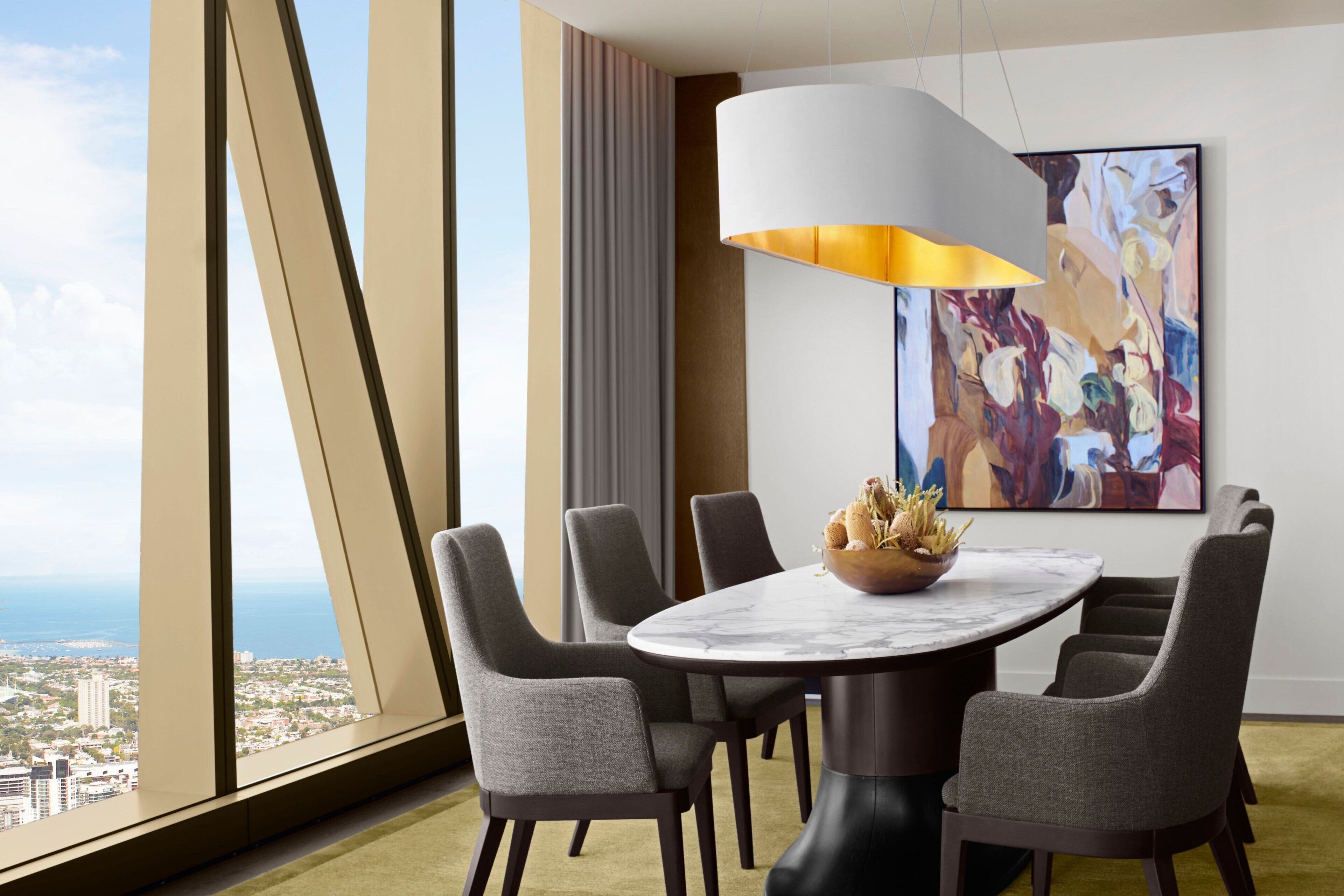 A modern dining area with a round table, six grey upholstered chairs, a marble top, and a large abstract painting on the wall.