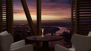 Een kamer met uitzicht op een stadsgezicht bij zonsondergang, een ronde tafel met een glas en twee stoelen.