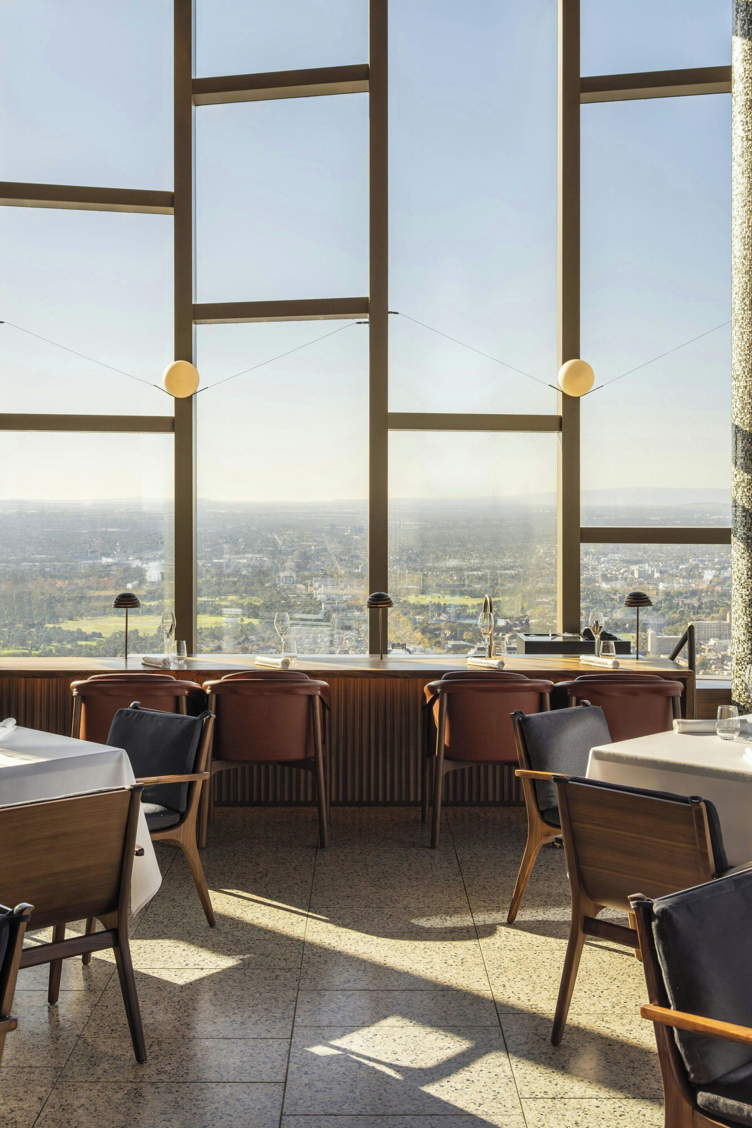 Un restaurante con amplios ventanales que ofrecen vistas a la ciudad. Las mesas están dispuestas con sillas, y hay lámparas colgantes.