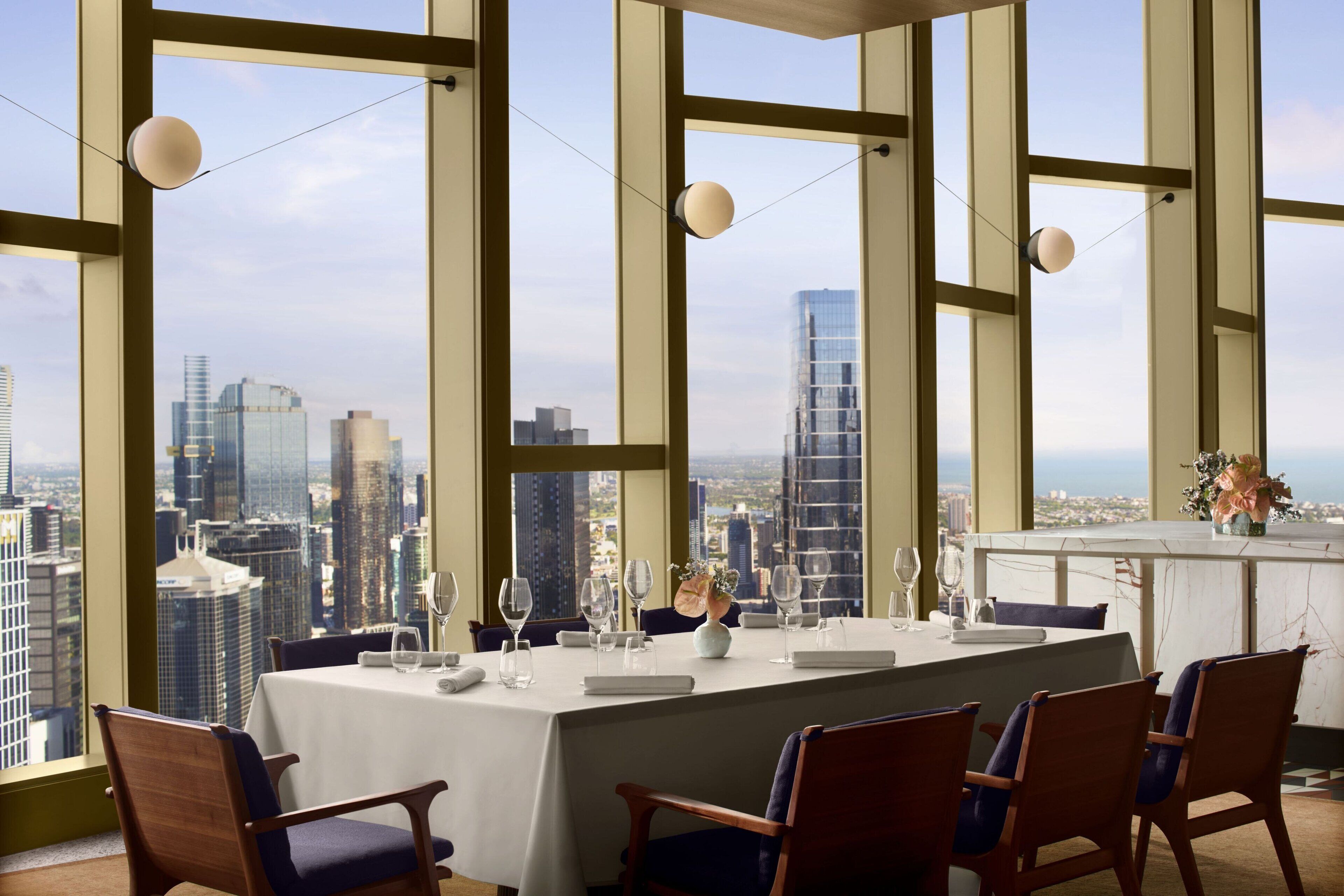 Comedor con mesa preparada para cuatro, ventanas amplias que ofrecen vistas a la ciudad e iluminación colgante.