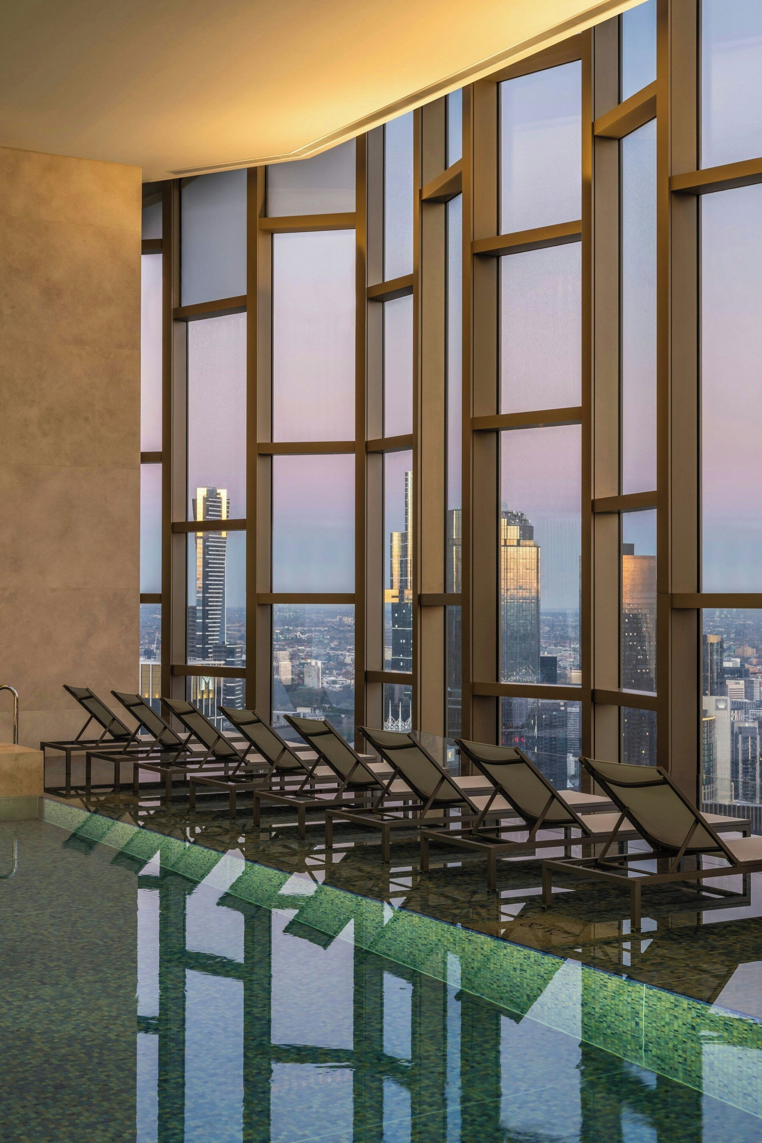 Un espace piscine intérieur moderne, doté de chaises longues et de grandes fenêtres offrant une vue sur la ville.