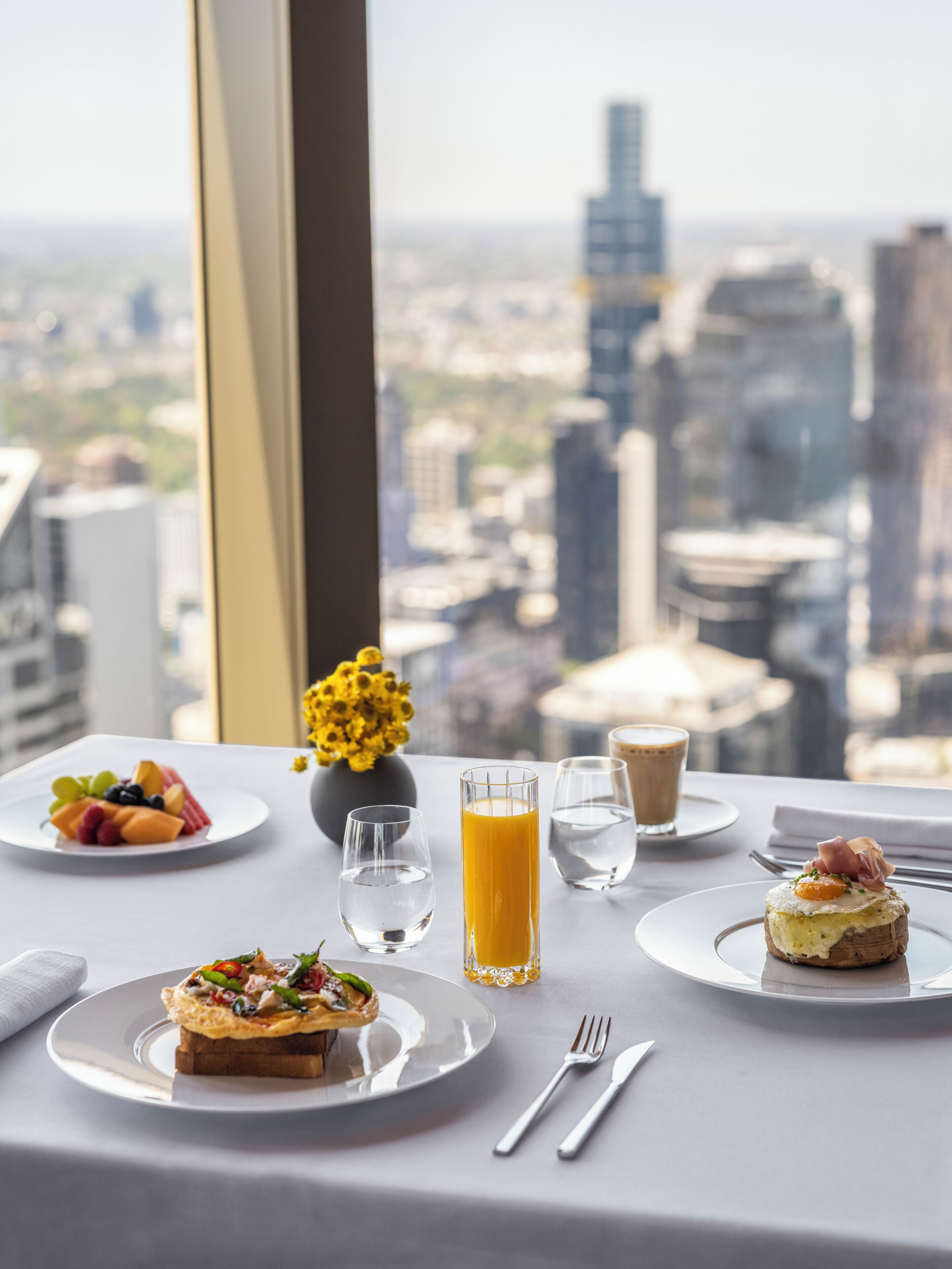 A table with breakfast items, a glass of orange juice, and a view of a cityscape through a window.