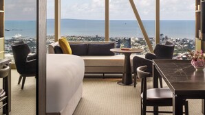 Una habitación de hotel moderna con un ventanal grande, un sofá, una mesa de comedor y vistas a la ciudad y al mar.