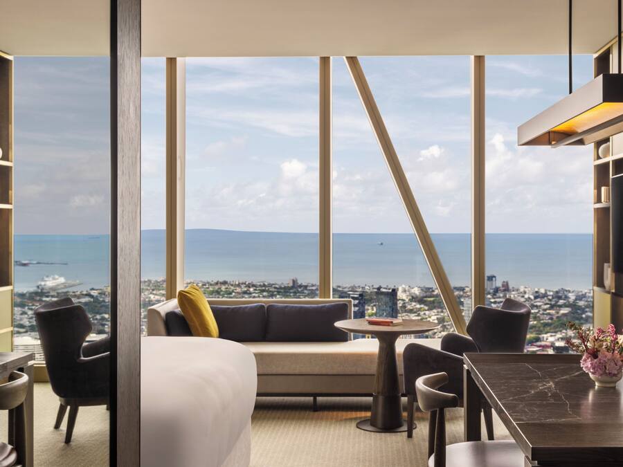 Una habitación de hotel moderna con un ventanal grande, un sofá, una mesa de comedor y vistas a la ciudad y al mar.