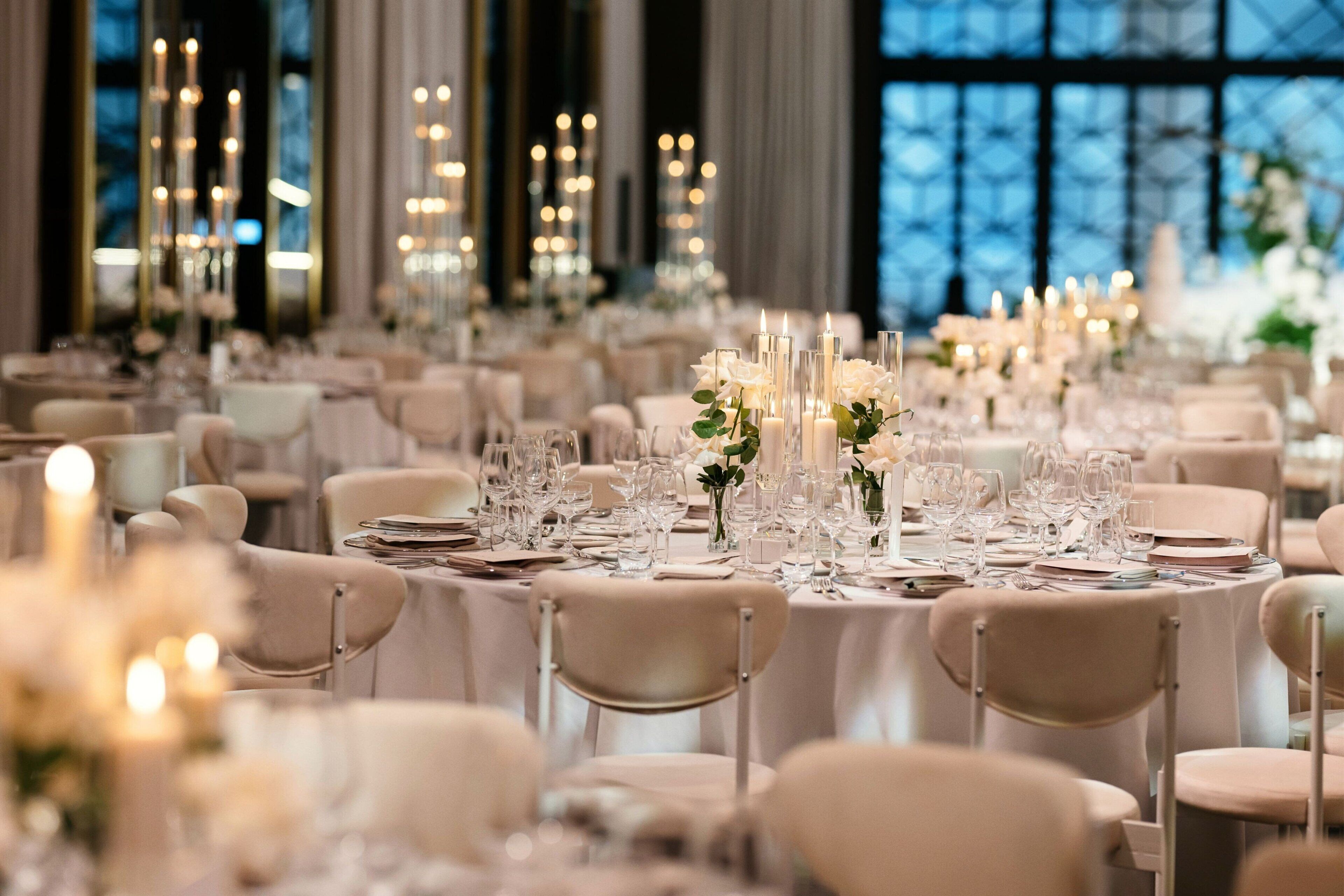 A banquet hall with round tables set for a formal event, featuring tall candles and floral centerpieces.