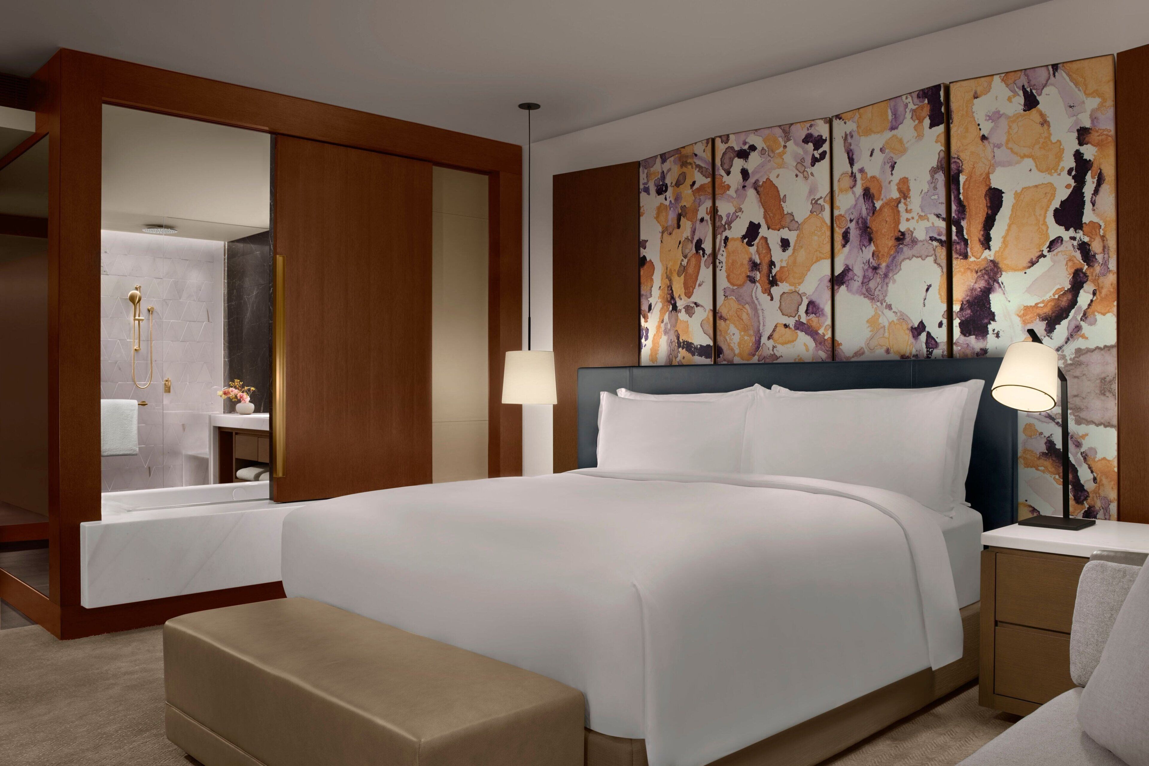 A modern hotel room with a large bed, a bedside table, a nightstand, a bench, a large mirror, and a wall with abstract art.