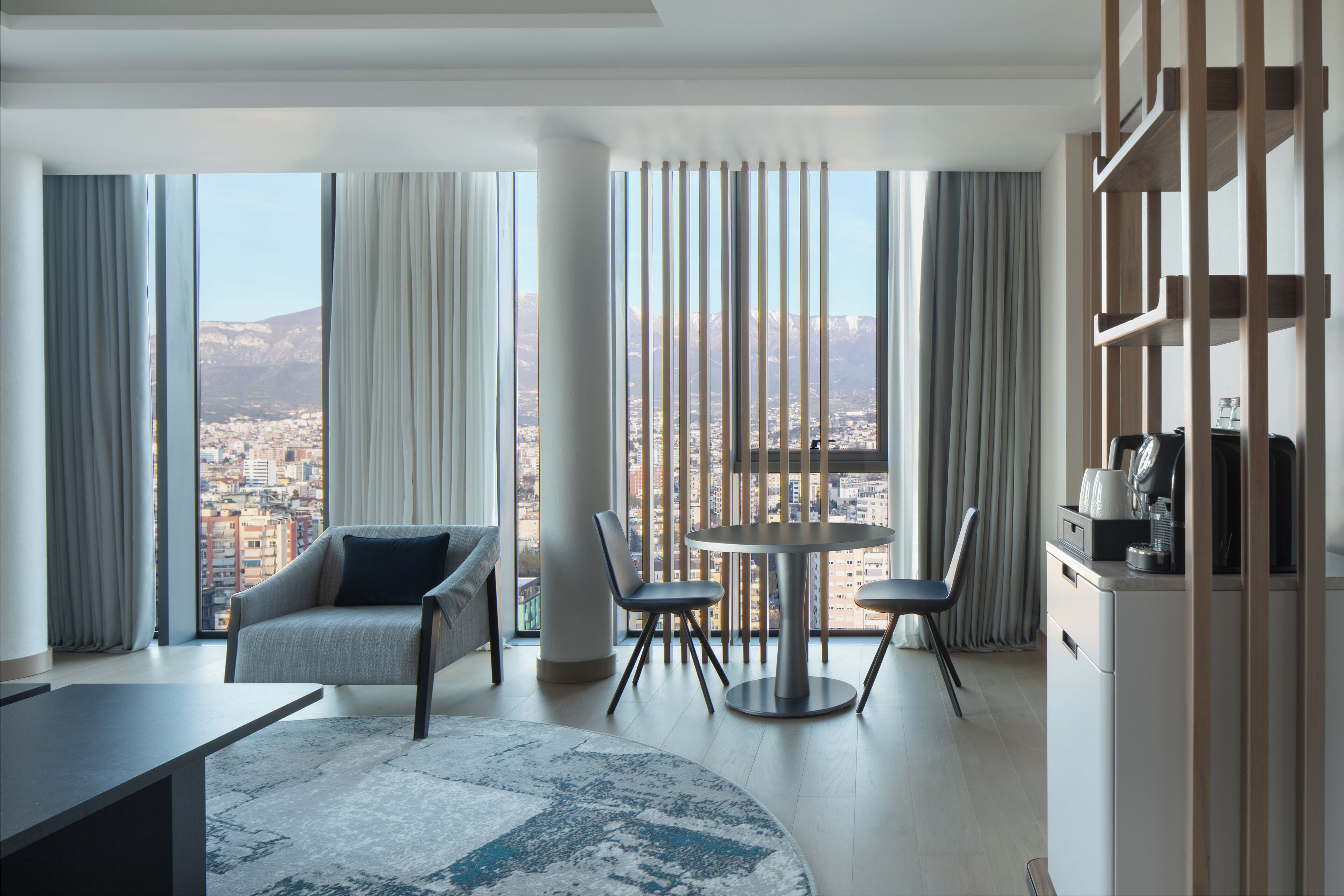 A modern hotel room with a large window offering a city view, a round dining table with chairs, a sofa, and a small kitchenette.