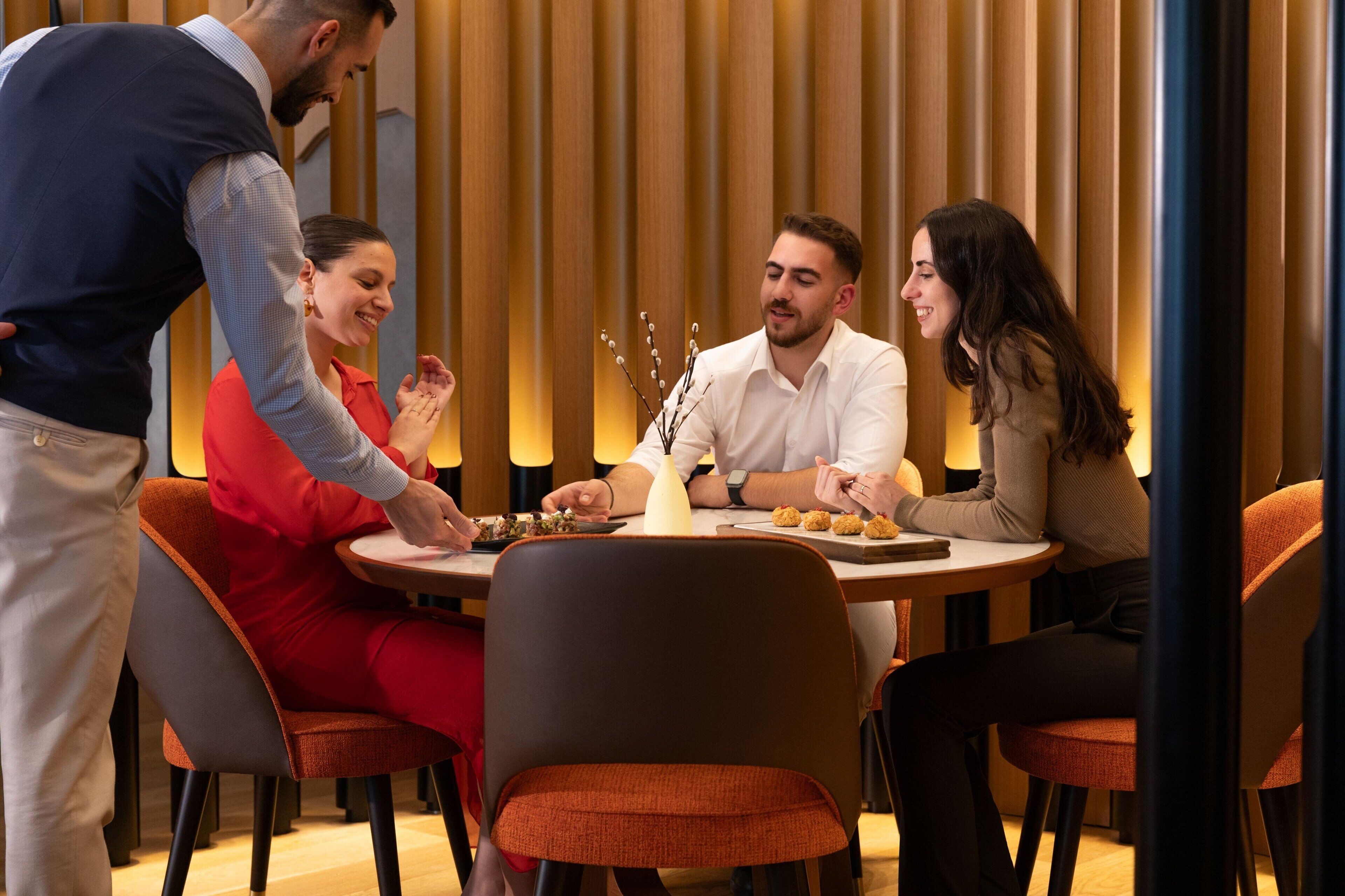 A group of people seated around a round table with a waiter standing beside them.