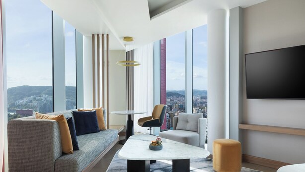 A modern hotel room with a large window offering a city view, a flat-screen TV mounted on the wall, a sofa, a chair, and a small coffee table with a vase of flowers. - Tirana Marriott (Tirana)