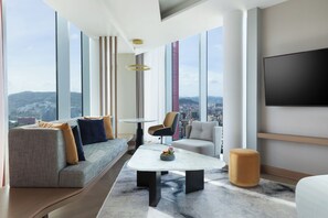 A modern hotel room with a large window offering a city view, a flat-screen TV mounted on the wall, a sofa, a chair, and a small coffee table with a vase of flowers. - Tirana Marriott (Tirana)