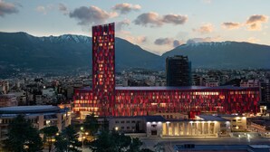 A modern building with a red facade and a tall tower, surrounded by a cityscape and mountains. - Tirana Marriott (Tirana)