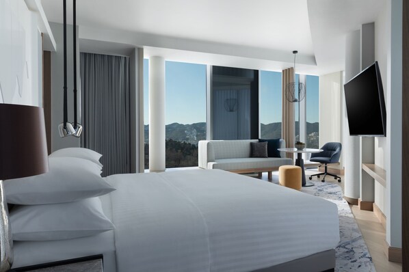 A modern hotel room with a large bed, a sofa, a TV, and a view of the mountains.