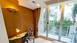 A hotel room with a bed, a small table with a lamp, a chair, and a large window with a view of greenery and buildings. - 2D - 3 Bedroom Downtown Apartment With Shared Pool (Bangkok)