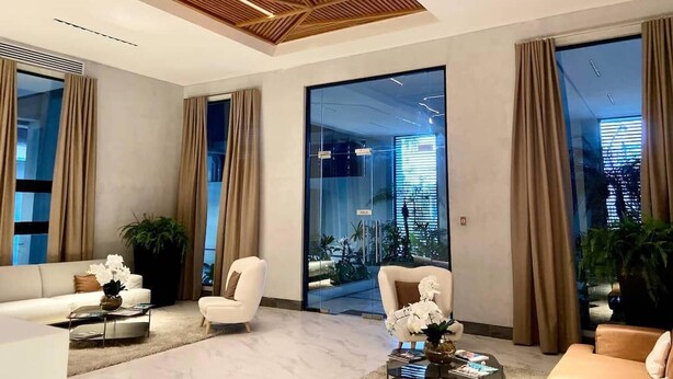 A modern lobby with a geometric ceiling design, large glass doors leading to a balcony, and a seating area with white armchairs and a coffee table. - Luxury Condo 1 Br Pool Excellent Location (Santo Domingo)