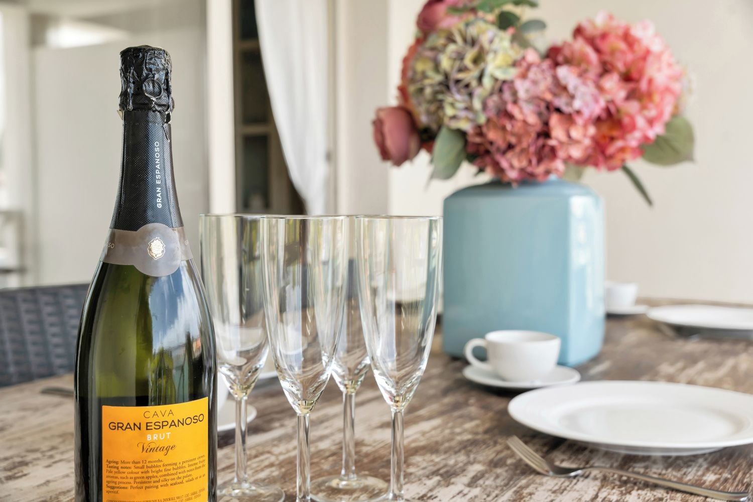 Uma garrafa de Cava Gran Espana Brut Vintage, quatro copos de vinho vazios, um vaso com flores, uma chávena branca e um prato sobre uma mesa de madeira.