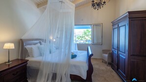 Un dormitorio con cama a dosel, mesita de noche, cómoda y ventana con vista a palmeras.