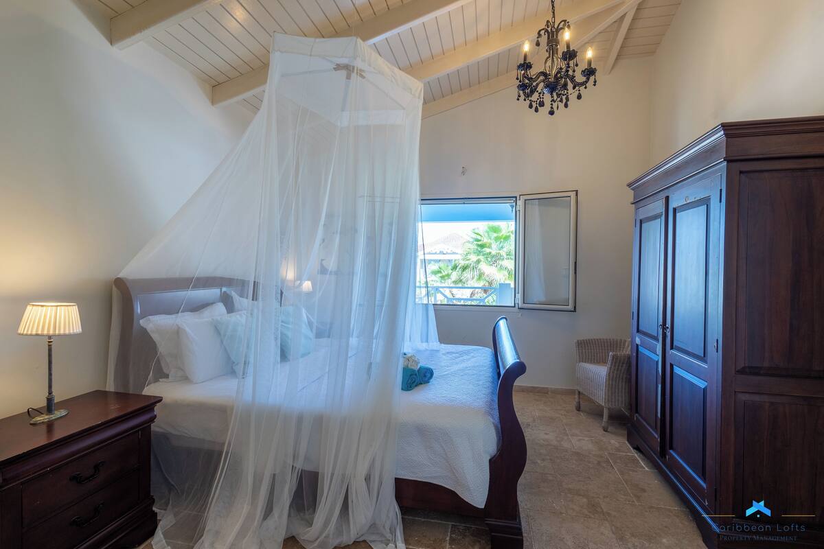 Ein Schlafzimmer mit Himmelsbett, Nachttisch, Kommode und einem Fenster mit Blick auf Palmen.
