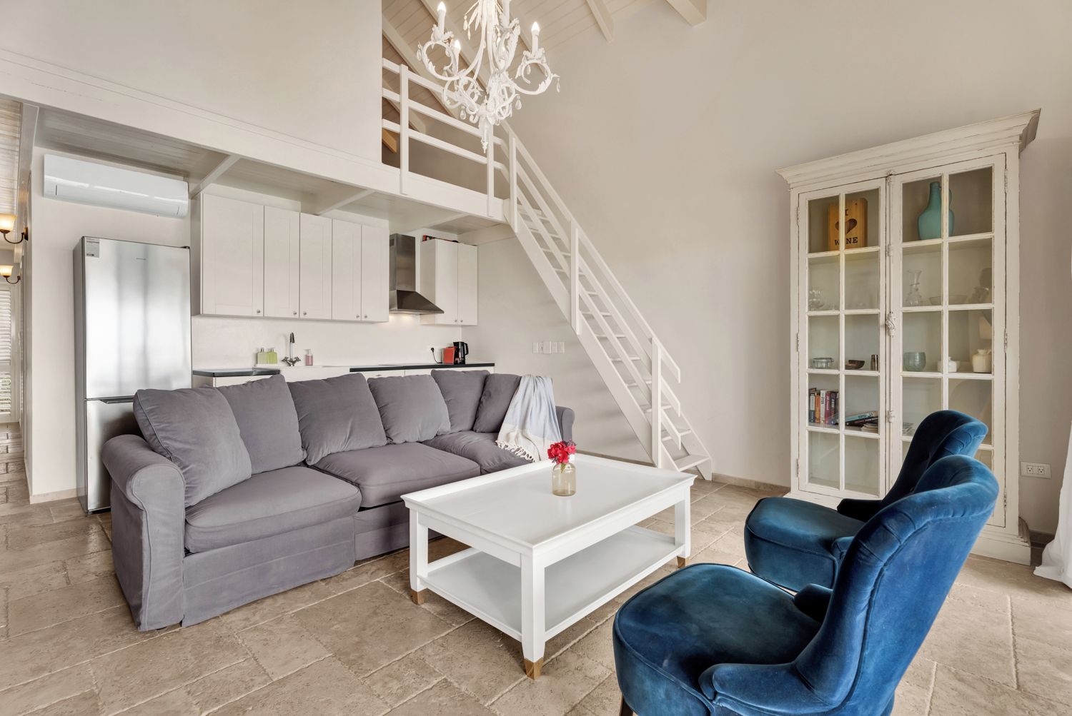 Una sala moderna con un sofá seccional gris, una mesa de centro blanca y un sillón azul. Una escalera blanca conduce a un nivel superior.