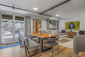 A modern living room with a wooden dining table, grey upholstered chairs, a sofa, and a large window with a view of a parking area. - No stairs required- 15 ppl unit in Camore, w/ Mountain View (Canmore)