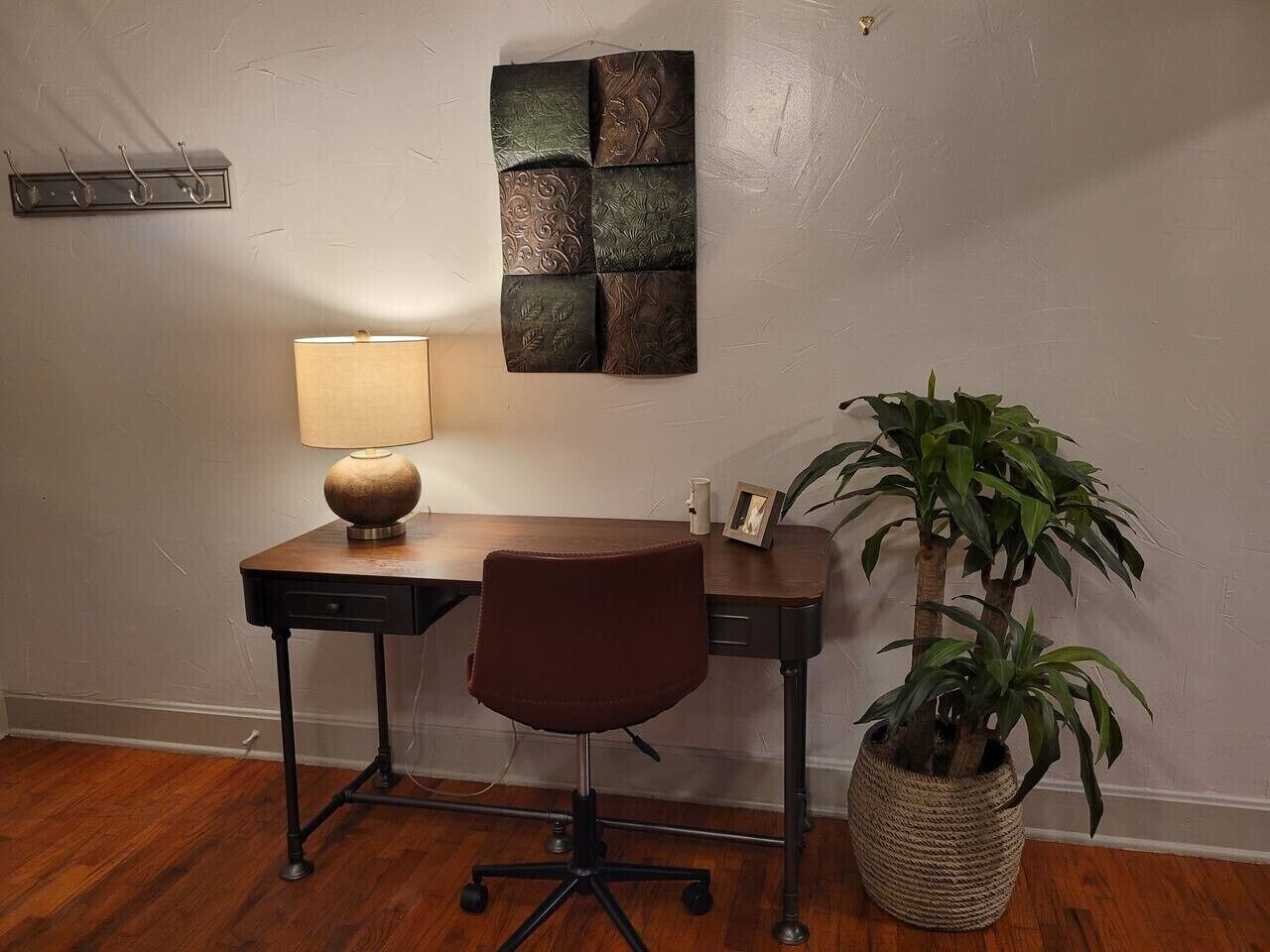 Uma mesa com luminária, uma cadeira e um vaso de planta em um quarto com piso de madeira e parede texturizada.