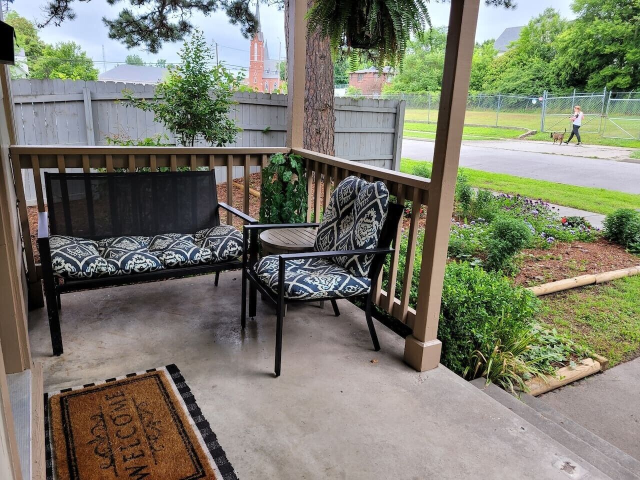 Een overdekt terras met een bankje en een stoel, een deurmat, en een tuin met planten en een hek.