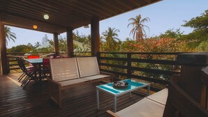 Eine Holzterrasse mit einer Couch und Stühlen, einem kleinen Tisch mit einer Schale sowie Blick auf Palmen und Grünpflanzen.