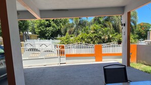 Ein Außenbereich mit Terrasse, zwei Stühle, ein Tisch, ein weißes Tor mit orangefarbenen Akzenten sowie Blick auf eine Straße mit Palmen und einem Gebäude im Hintergrund.