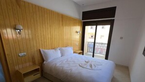 A bedroom with a wooden panel wall, a bed with white bedding, a small wooden nightstand, a wall-mounted light, and a view of the outside balcony. - Appartment with Moroccan breakfast (Dakhla)