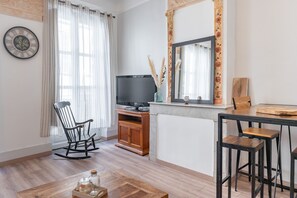 A living room with a wooden rocking chair, a TV on a stand, a dining table with stools, and a large mirror above the mirror.