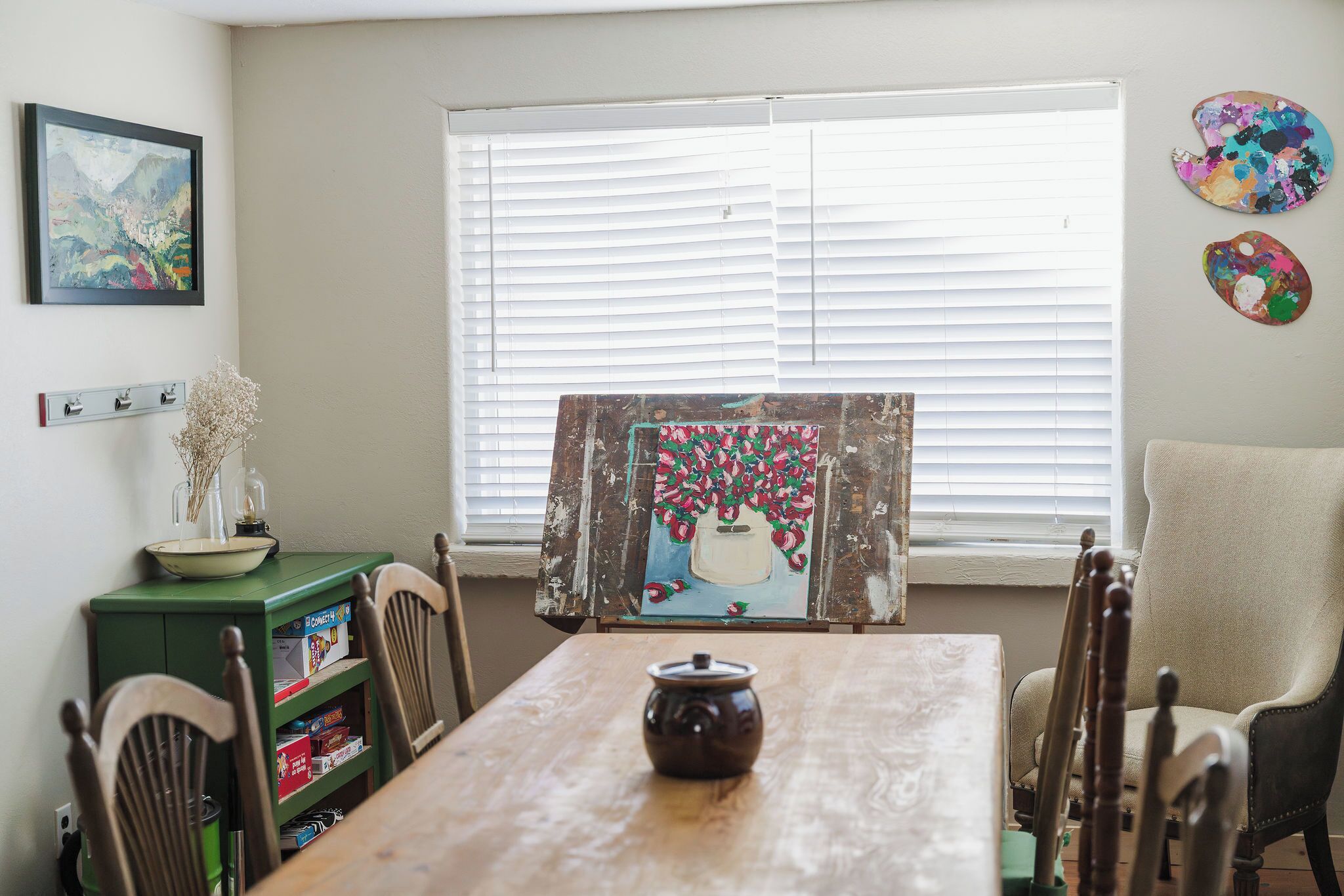 A dining area with a wooden table, chairs, a green cabinet with board games, a painting on the wall, and a window with blinds.